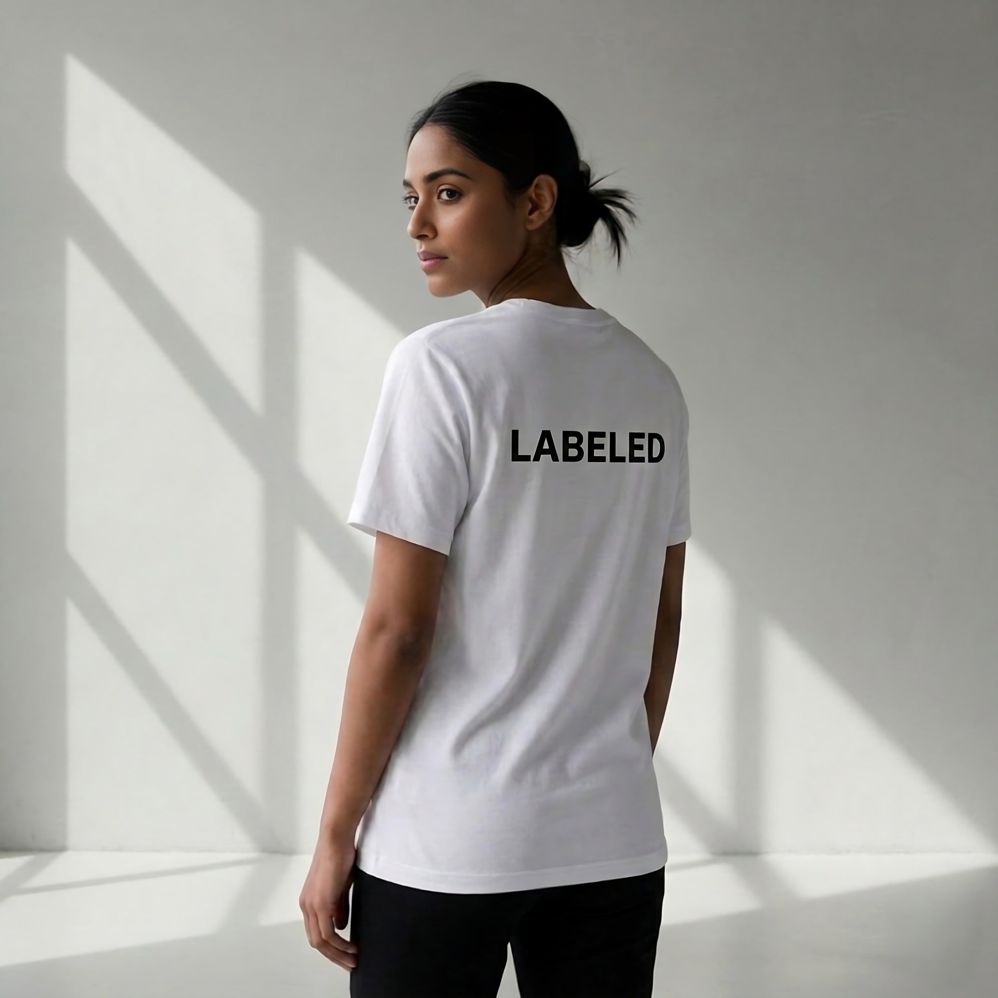 Woman wearing white t-shirt with 'LABELED' text on the back in a minimalistic setting.